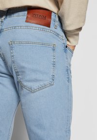 Šviesiai mėlyni džinsai pagaminti iš džinso audinio, ant nugaros yra rudas odinis lopas su iškaltu tekstu. Dizainą sudaro standartinės nugaros kišenės su subtiliu siuvimu.