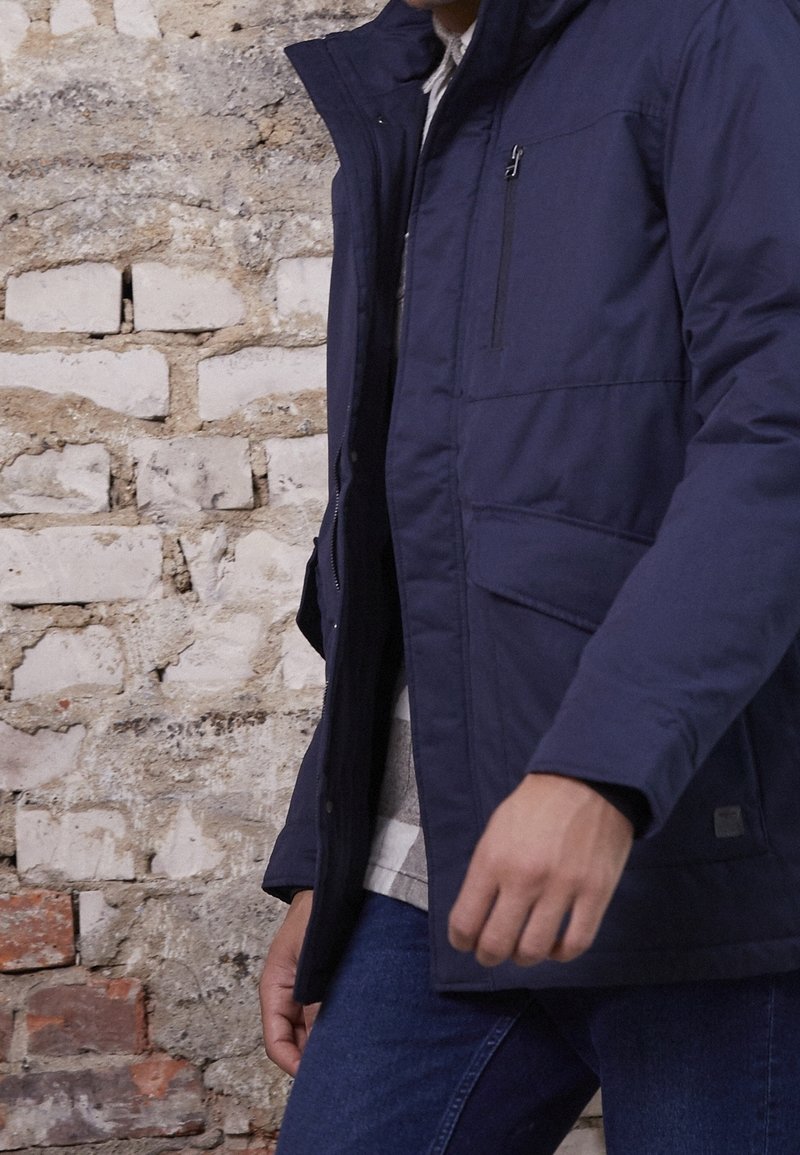 Personne portant une veste bleu marine avec une poche poitrine zippée et une poche à rabat, debout contre un mur en briques blanc et rouge.