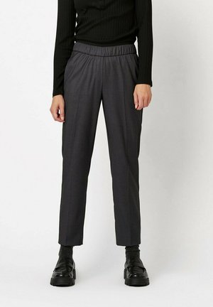 Person trägt ein schwarzes Langarmoberteil, dunkelgraue elastische Bundhose, schwarze Socken und schwarze Loafers und steht vor weißem Hintergrund.