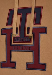 Béžová mikina s kapucí, zdobená vyšitým logem v tmavě modré a vínové barvě, s tkaničkami a zlatými akcenty s nápisem "TOMMY HILFIGER."