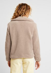 Beige fleecejakke med krave, lange ærmer og et blødt, tekstureret stof. Båret over lysegule bukser.