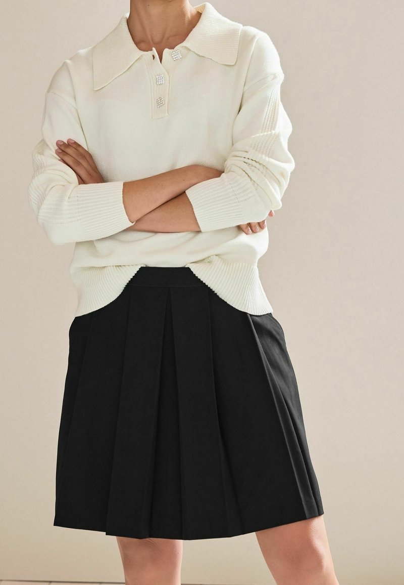 Weißer Strickpullover mit Kragen und dekorativen Knöpfen, kombiniert mit einem schwarzen, plissierten Rock. Beide Kleidungsstücke haben eine glatte Textur.