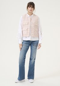 Gilet in tweed rosa con due tasche frontali, camicia bianca con maniche legate e jeans blu a zampa d'elefante. Il modello si trova su uno sfondo neutro.