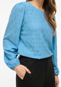 Hellblaue, strukturierte Bluse mit einem floralen Lochstickmuster und langen Ärmeln, kombiniert mit schwarzen, maßgeschneiderten Hosen; elastische Manschetten an den Ärmeln.