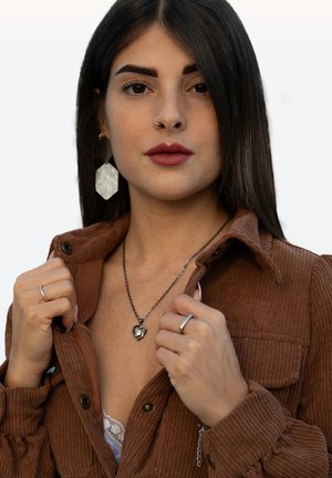 Brown corduroy jacket with button details, layered over a white lace top. Silver heart necklace, large hexagonal earrings, and rings are visible.