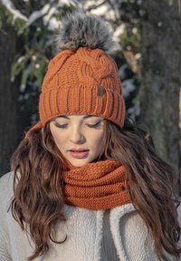 Berretto lavorato a maglia arancione con un pompon grigio soffice e design a trecce, abbinato a una sciarpa arancione a costine, posizionato su uno sfondo innevato.