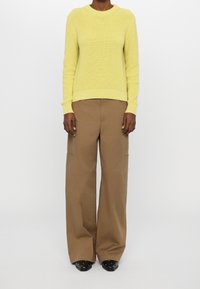 Maglione giallo a coste, con collo a giro, abbinato a pantaloni larghi marroni e scarpe nere. L'outfit presenta una vestibilità rilassata e dettagli minimi.