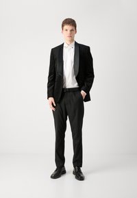 Svart tuxedojacka med texturerad tyg, satinrevär, vit skjorta, svarta byxor och svarta skor, visad på en modell.