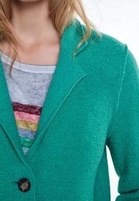 Blazer en laine teal avec revers crantés, fermeture à un bouton, et tissu texturé, superposé à un pull gris à rayures multicolores.