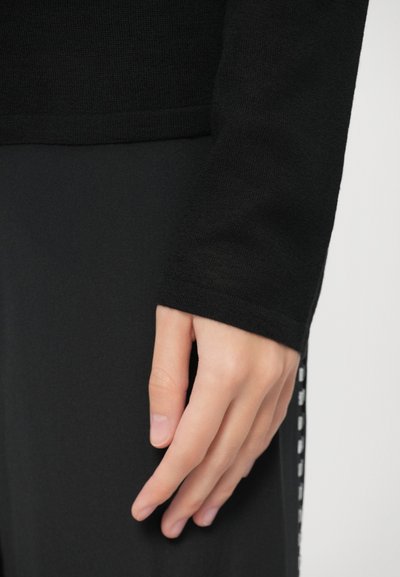 Mustast soonikootud pikkade varrukatega topp, millel on kitsad varrukaotsad, kombineeritud mustade pükstega, mille küljes on vertikaalne valge mustriline triip.