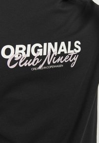 Camiseta de algodón negra con un texto blanco audaz: "ORIGINALS Club Ninety" y "CREATED IN COPENHAGEN" en un tamaño de fuente más pequeño, sobre un fondo de textura suave.