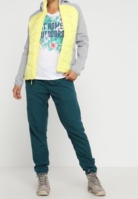 Ljusgrå och gul quiltad jacka över en vit blommig grafisk t-shirt, tillsammans med teal joggers och beige vandringskängor.