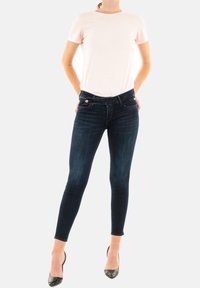 Jean skinny en denim foncé avec un léger brillant, doté d'un design classique, de poches avant et de matériel doré contrastant. Associé à un t-shirt rose clair.