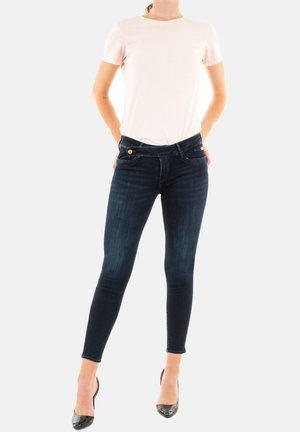 Jean skinny en denim foncé avec un léger brillant, doté d'un design classique, de poches avant et de matériel doré contrastant. Associé à un t-shirt rose clair.