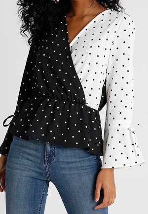 Femme portant un chemisier cache-cœur à pois noirs et blancs avec une taille volantée, assorti à un jean bleu.