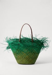 Bolso cesta verde tejido con detalle de flecos, con asas de cuero marrón. Superficie texturizada con un degradado de verde oscuro a claro.