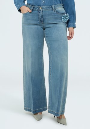 Donna che indossa jeans a gamba larga color azzurro chiaro con un dettaglio floreale vicino alla tasca e tacchi grigi con punta affilata, in piedi con una mano in tasca.