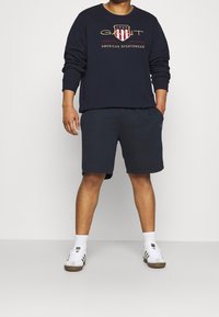 Felpa della marina con design del logo, abbinata a pantaloni corti scuri. Sneakers bianche con dettagli marroni e calze bianche. Abbigliamento sportivo casual.