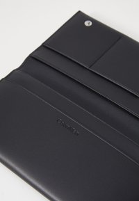 Čierna kožená peňaženka s viacerými priehradkami na karty, textúrovaným povrchom a strieborným zapínaním na patent. Na prednej strane sa nachádza embosované logo.
