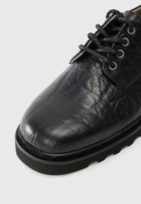 Pantof de piele neagră cu o suprafață texturată, vârf rotund, șireturi negre și ochiuri metalice. Dispune de o talpă groasă din cauciuc negru.