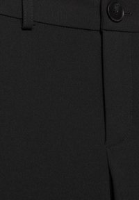 Lähikuva mustista kangashousuista, joissa näkyy vyötärönauha, vyölenkki, nappi ja taskun sauman yksityiskohdat.