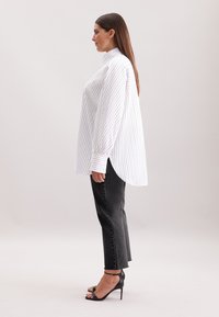 Camicia bianca a righe con colletto alto, maniche lunghe e spacchi laterali, abbinata a jeans grigi scuri a gamba dritta e sandali neri con tacco alto.