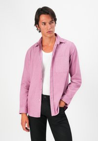 Young man wearing a pink corduroy shirt over a white tank top and black pants, standing with one hand in pocket, looking to the side.