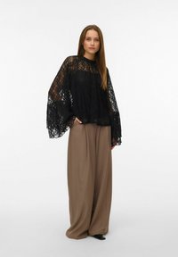 Svart bluse i blonder med vide, flagrende ermer kombinert med høytlivede, vide brune bukser. Antrekket kombinerer teksturert stoff og avslappet silhuett.