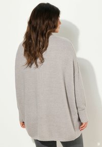 Světle šedý oversized pletený svetr s žebrovanými manžetami a lemem. Má uvolněný střih a strukturovaný vzor. Stylizováno s tmavě šedými kalhotami.