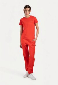 Rood atletisch shirt en bijpassende broek van gladde stof. Het shirt heeft een klein logo en de broek heeft zijzakken en een ontspannen pasvorm.