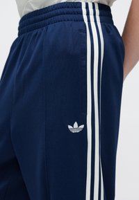 Mornarsko plave sportske hlače s bijelim trakastim detaljima u trima linijama, elastičnim pojasom i malim bijelim logotipom na donjem dijelu nogavice. Glatka tekstura tkanine.