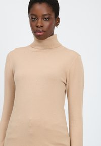 Senza colletto beige realizzato in morbido tessuto a maglia fine. Design aderente con maniche lunghe e colletto a coste. Texture liscia, dettagli minimi.