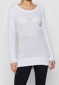 Pull blanc en maille avec un design ouvert et lâche, col rond large, manches longues et ourlet côtelé. Tissu texturé avec légère transparence.