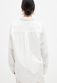 White cotton shirt with a back yoke, long sleeves, and buttoned cuffs. Features a classic collar and a relaxed fit with a pleat detail.