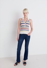 Adīta halter top ar daudzkrāsainām svītrām, baltu pamatu un ar rāvējslēdzēju; apvienots ar tumši zilām platām džinsiem un melniem sandālēm ar koka papēžiem.