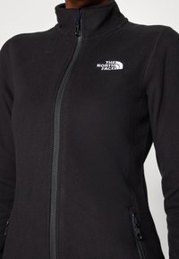 Zwarte fleece jack met hoge kraag, rits aan de voorkant en zijzakken. Voorzien van een geborduurd logo op de linkerborst en een zachte textuur.