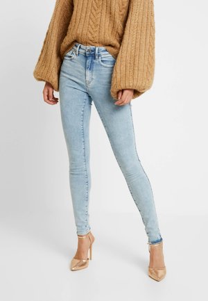 Personne portant un jean skinny bleu clair, un pull en maille surdimensionné camel et des chaussures à talons hauts dorées, debout devant un fond uni.