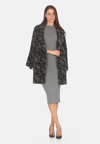 Cappotto in lana a motivi neri e grigi con ampio colletto e design doppiopetto, indossato sopra un vestito grigio attillato e tacchi neri.