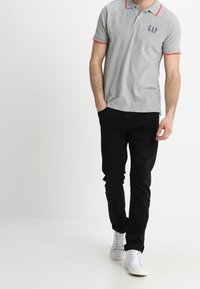 Polo gris avec accents de col rouge et logo "GAP" bleu marine, associé à un pantalon noir et des baskets blanches. Modèle se tenant contre un fond blanc.