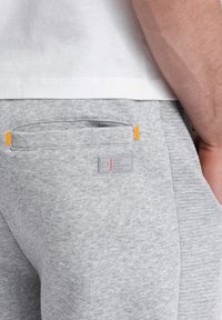 Graue Sweatpants aus einem strukturierten Stoff, mit einer Seitentasche in Orange-Akzenten und einem Label-Patch in der Nähe des Bundes.