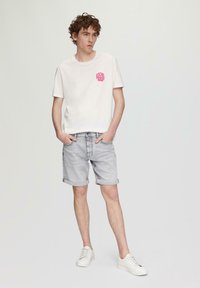 Weißes T-Shirt mit pinkem kreisförmigem Grafikdesign, beigen Denim-Shorts mit aufgerollten Säumen und weißen Sneakers; einfarbiger Hintergrund.