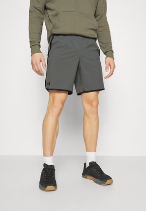 Mand iført grå sportsshorts, olivengrøn hættetrøje, hvide sokker og sorte sportssko står foran en ensfarvet baggrund.