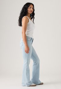 Hellblaue Schlagjeans mit mittelhohem Bund aus Denim, kombiniert mit einem weißen, gerippten Tanktop; Modell steht im Profil vor neutralem Hintergrund.