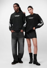 Hoodies pretas com desenhos de chamas brancas, emparelhados com calças largas cinzas e uma saia de couro preta, complementados por botas de tornozelo pretas.