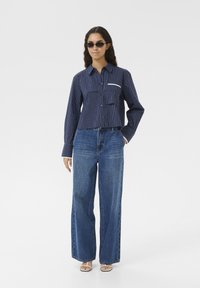 Marine gestreepte cropped shirt met twee voorzakken, gecombineerd met wijde blauwe jeans. Model draagt donkere zonnebril en open teen sandalen.