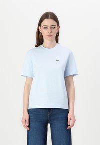 Camiseta de algodón azul claro con manga corta, cuello redondo y un pequeño logotipo de cocodrilo verde en el lado izquierdo del pecho. Ajuste casual, textura suave.