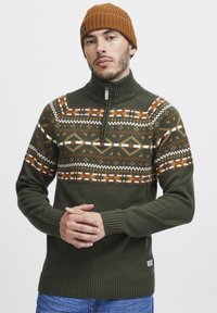 Pulover din tricot olive cu guler cu fermoar, având un model pe piept în nuanțe de portocaliu, alb și gri închis. Mânecile și tivul sunt ribbate.