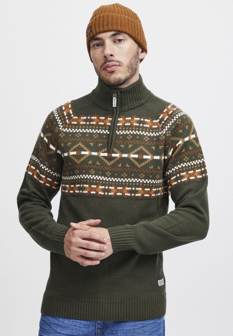 Sweter w oliwkowym zielonym kolorze, dziany, z zamkiem przy szyi, z wzorzystą klatką piersiową w odcieniach pomarańczowym, białym i ciemnoszarym. Rękawy i dół wykończone ściągaczem.