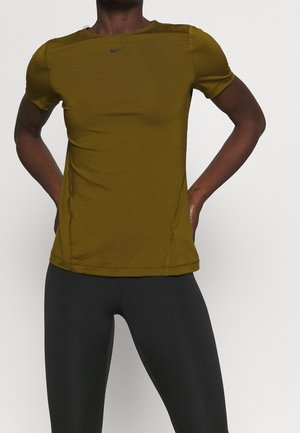 Mand iført olivengrøn kortærmet sportstrøje og sorte leggings, der står med hænderne på hoften mod en ensfarvet baggrund.