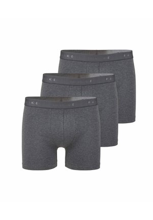 Athletischer Mann steht barfuß da und trägt dunkelgraue Boxershorts vor einem einfarbigen hellen Hintergrund.